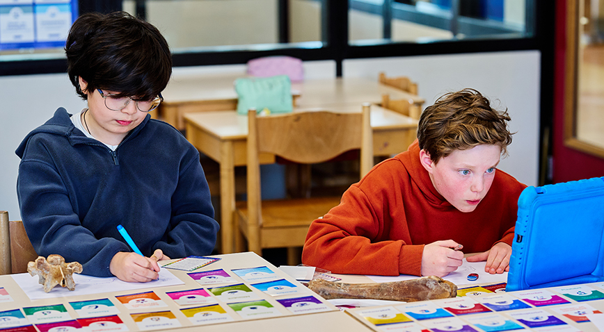 Twee montessori-leerlingen zitten ingespannen te werken aan een tafeltje; een van hen heeft een blauwe tablet voor zich en op tafel liggen allemaal gekleurde kaartjes en twee botten
