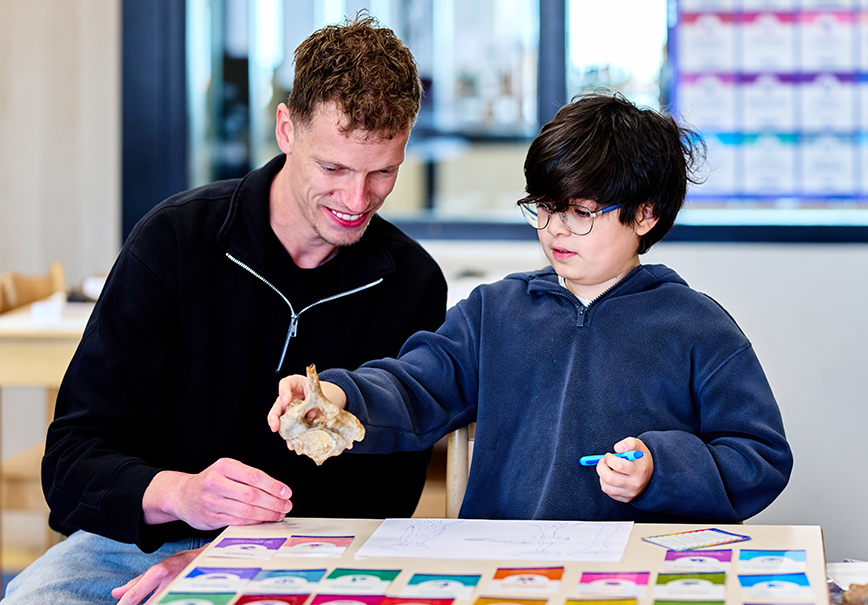 Een montessori-leerkracht zit met een leerling bij een tafeltje; de leerling heeft een bot in zijn hand en op tafel liggen allemaal kaartjes, ook ligt er een tekening van de leerling