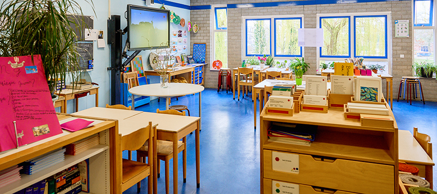 Montessori klaslokaal van een onderbouw groep met tafeltjes, kasten, plantjes en een digitaal onderwijsbord