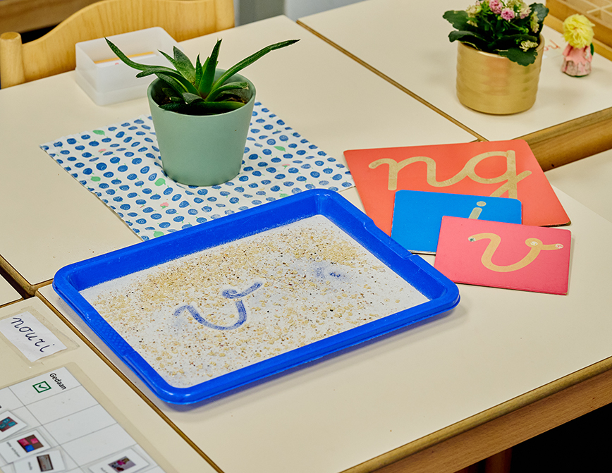 Montessori-lesmateriaal om te leren schrijven; op een tafel ligt een dienblad met zand waar de letter v in is geschreven; daarnaast liggen bordjes met letters van schuurpapier