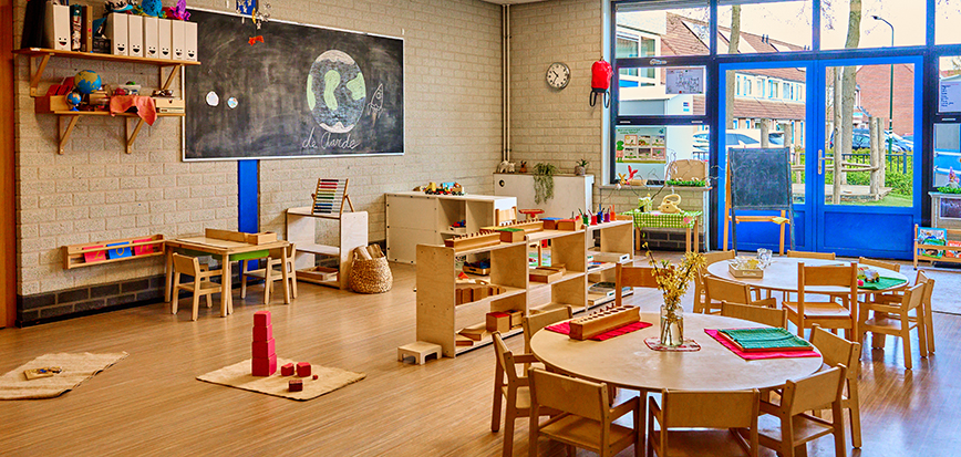 Montessori-klaslokaal van een peutergroep met ronde tafeltjes en stoeltjes er omheen; op de grond liggen montessori-werkjes en er is een groot schoolbord