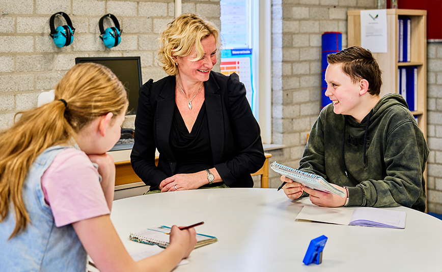 Een montesssori-leerkracht zit met twee leerlingen aan tafel; op de tafel liggen werkjes