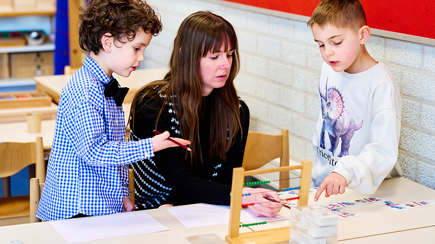 Twee jonge montessori-leerlingen laten aan hun leerkracht zien dat zij de stof beheersen; op tafel staat een telraam en liggen kaartjes